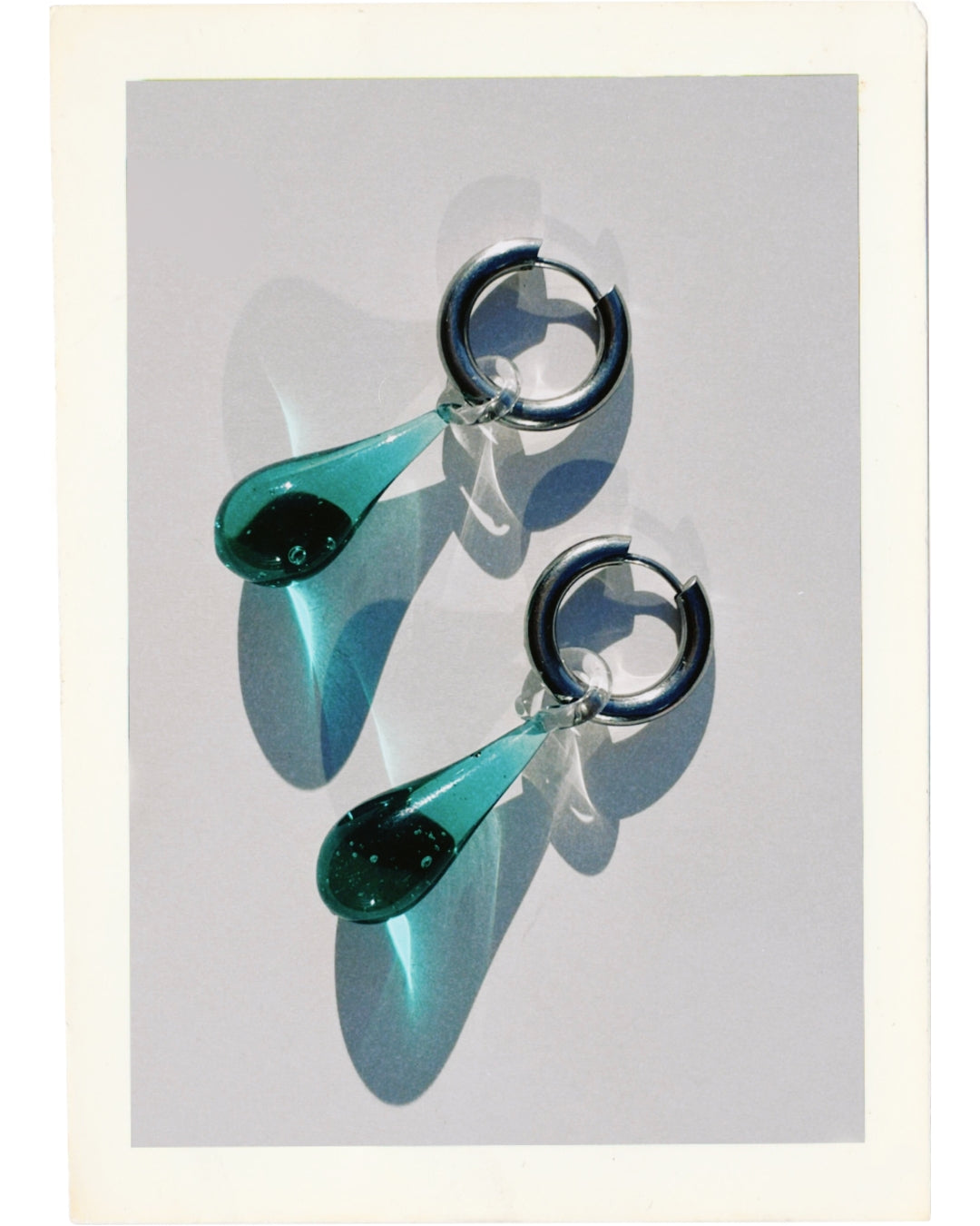 A close-up studio shot of The Celeste Earrings is made with thick Sterling Silver/18k gold plated tarnish-resistant, hypoallergenic 12 mm thick hoops and high-quality 22mm lampwork glass blue water drop-shaped pendants.