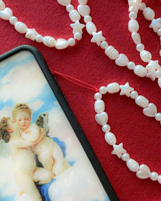 A close up studio shot on red felt of multiple Buttercup Studio's Love Is In The Air Phone Straps. Made with white freshwater pearls and white star and heart shaped beads. One phone strap is attached to a phone case featuring two winged human cherubs.