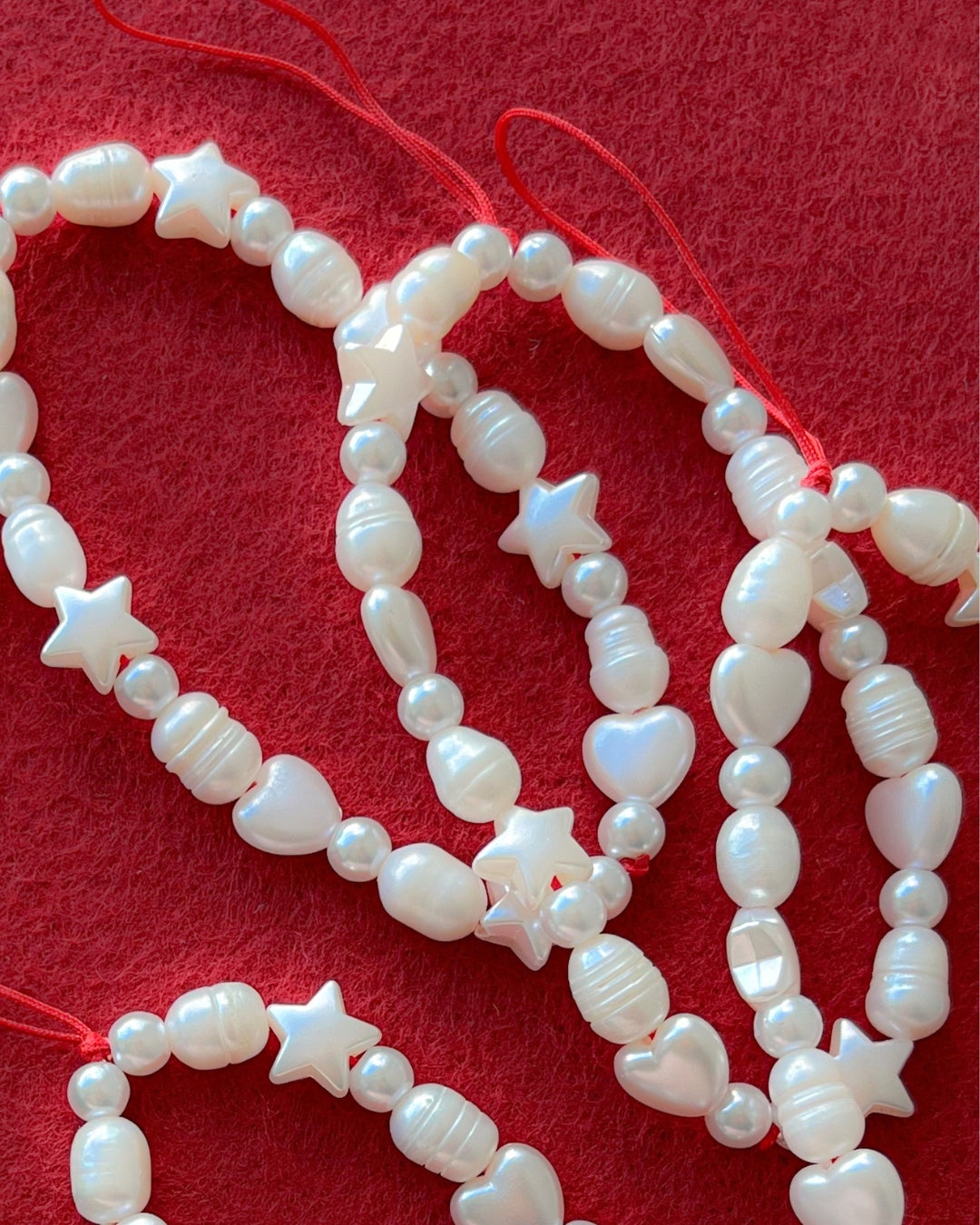 A close up studio shot on red felt of multiple Buttercup Studio's Love Is In The Air Phone Straps. Made with white freshwater pearls and white star and heart shaped beads.