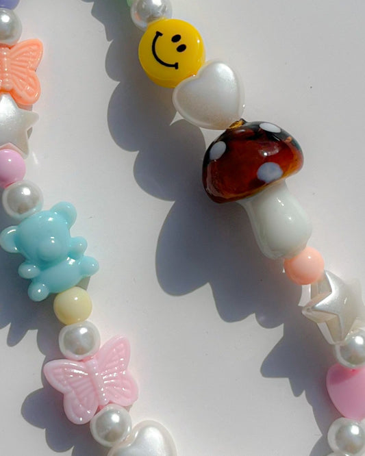 A close up studio shot of Buttercup Studio's Mushroom Forest Wonderland Phone Strap. Made with pearls, white star and heart beads, colourful beads, a blue bear bead, pink and orange butterfly beads, a smiley face bead and a special brown mushroom lampwork glass bead.