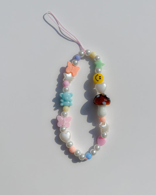A studio shot of Buttercup Studio's Mushroom Forest Wonderland Phone Strap. Made with pearls, white star and heart beads, colourful beads, a blue bear bead, pink and orange butterfly beads, a smiley face bead and a special brown mushroom lampwork glass bead. 