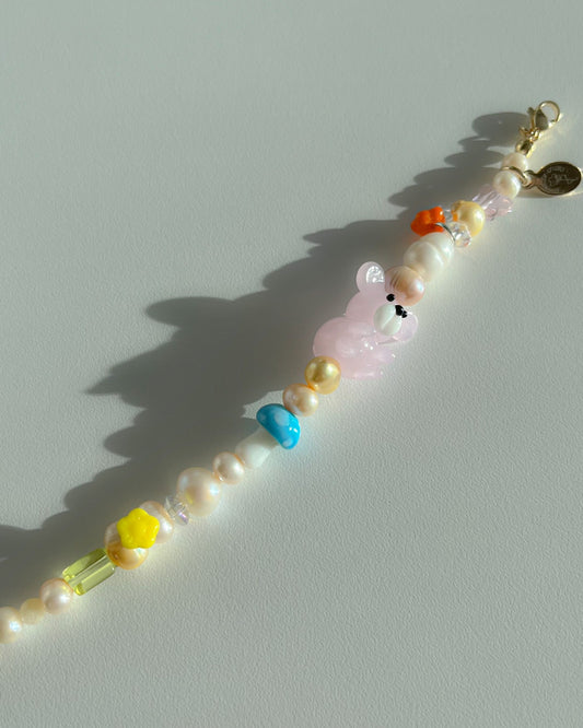 A studio shot of Buttercup Studio's Pink Forest Teddy Freshwater Pearls Bracelet. Made with assorted freshwater pearls, green, yellow, orange and pink charms, a special lampwork blue mushroom glass bead and a special pink teddy lampwork glass bead.