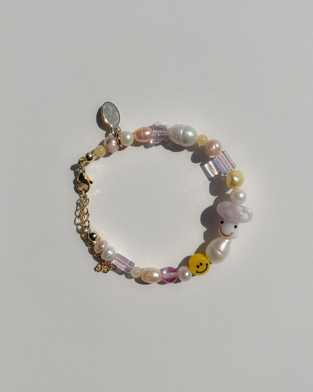 A studio shot of Buttercup Studio's Pink Shroomie Freshwater Pearls Bracelet. Includes assorted freshwater pearls, sheer pink beads, a yellow smiley face bead and a special lampwork glass pink mushroom bead.