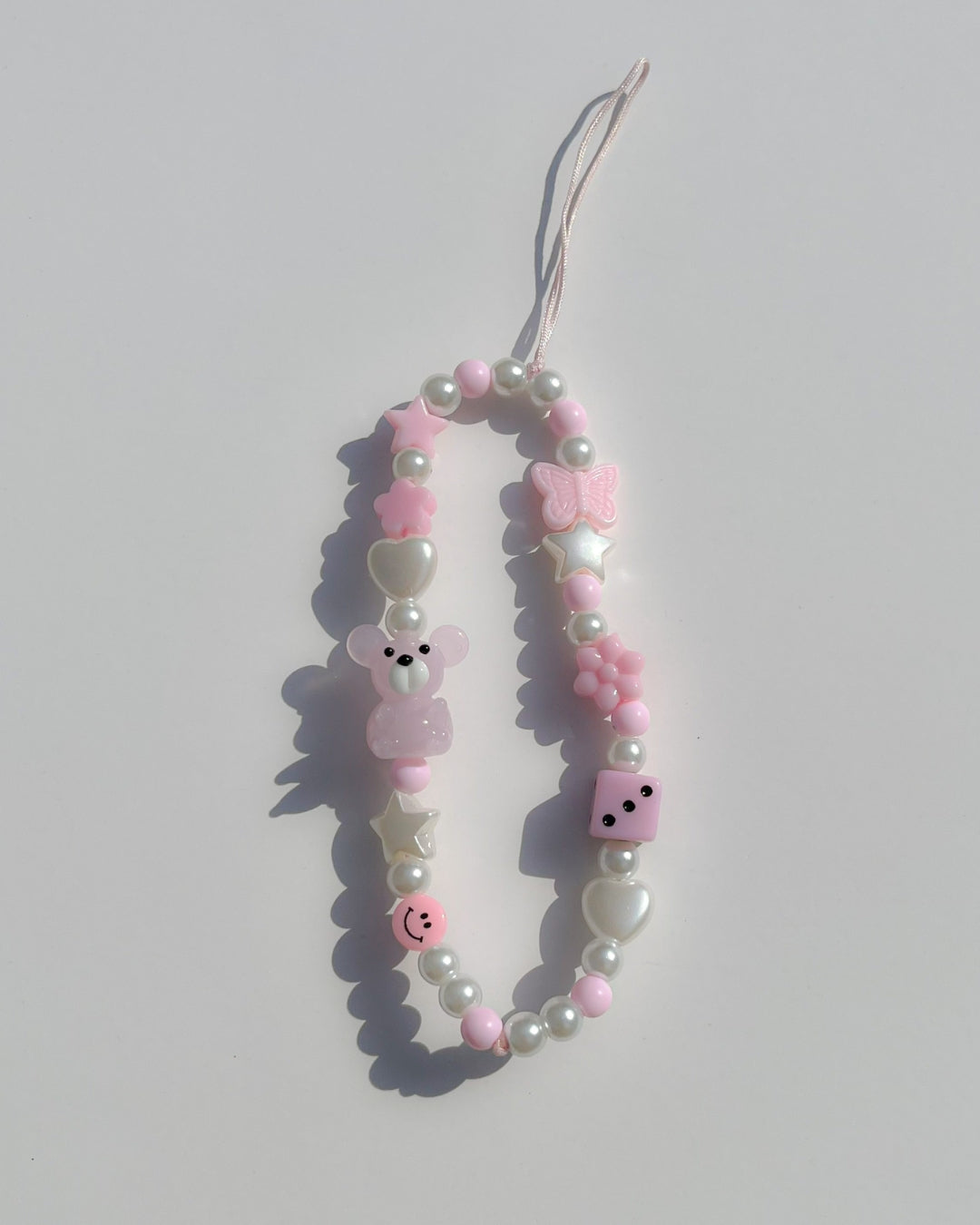 A studio shot of Buttercup Studio's Pink Teddy Princess Phone Strap. Made with pearls, white star and heart beads, a pink butterfly bead, a pink dice bead, a pink smiley face bead and a lampwork glass pink teddy bead.