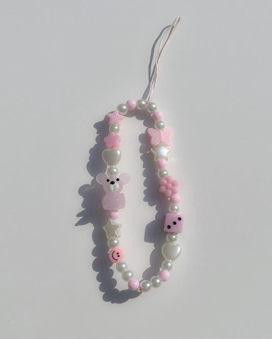 A studio shot of Buttercup Studio's Pink Teddy Princess Phone Strap. Made with pearls, white star and heart beads, a pink butterfly bead, a pink dice bead, a pink smiley face bead and a lampwork glass pink teddy bead. 