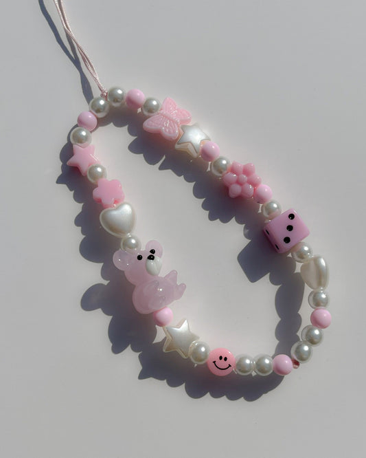 A studio shot of Buttercup Studio's Pink Teddy Princess Phone Strap. Made with pearls, white star and heart beads, a pink butterfly bead, a pink dice bead, a pink smiley face bead and a lampwork glass pink teddy bead.