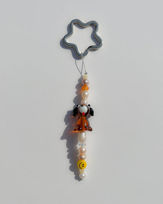 A studio shot of the one of one Puppy Love Freshwater Pearls Lucky Charm keychain. Handmade using stainless steel wire looped on a star shaped keyring, this freshwater pearl keychain features assorted freshwater pearls, a smiley face bead and a special lampwork brown puppy glass bead. 