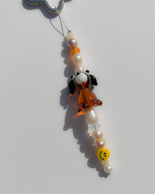 A studio shot of the one of one Puppy Love Freshwater Pearls Lucky Charm keychain. Handmade using stainless steel wire looped on a star shaped keyring, this freshwater pearl keychain features assorted freshwater pearls, a smiley face bead and a special lampwork brown puppy glass bead. 