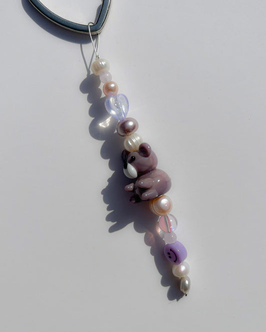 A studio shot of the one of one Purple Teddy Freshwater Pearls Lucky Charm keychain. Handmade using stainless steel wire looped on a heart shaped keyring, this freshwater pearl keychain features assorted freshwater pearls, a smiley face bead and a special lampwork purple teddy glass bead. 