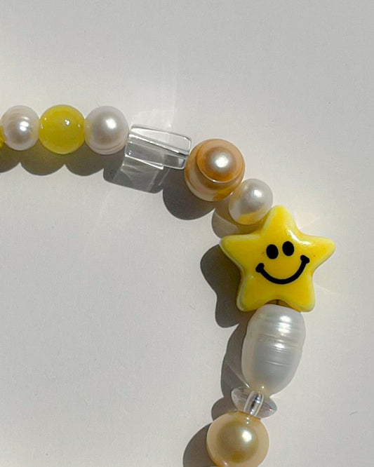 A close up studio shot of Buttercup Studio's Shooting Star Freshwater Pearls Bracelet. Made with assorted freshwater pearls, yellow and white beads, and a special lampwork glass smiley face star bead.