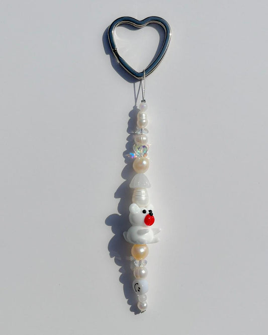 A studio shot of the one of one Snowy Teddy Freshwater Pearl Lucky Charm keychain. Handmade using stainless steel wire looped on a heart shaped keyring, this freshwater pearl keychain features assorted freshwater pearls, a smiley face bead and a special lampwork white bear with a red nose glass bead. 