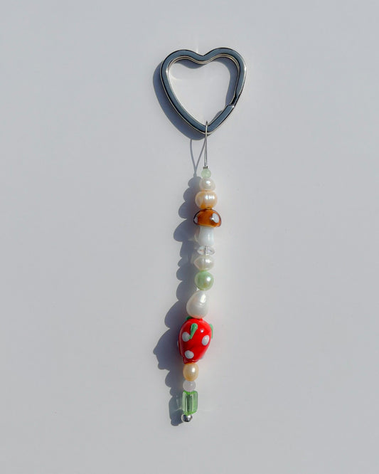 A studio shot of the one of one Strawberry Baby Freshwater Pearls Lucky Charm keychain. Handmade using stainless steel wire looped on a heart shaped keyring, this freshwater pearl keychain features assorted freshwater pearls, a lampwork glass brown mushroom bead and a lampwork glass strawberry bead.