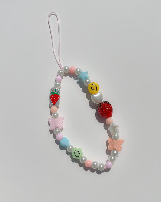 A studio shot of Buttercup Studio's Strawberry Jam Phone Strap. Made with pearls, colourful beads, white heart and star beads, a pink butterfly bead, two smiley face beads, a glass hand painted rectangle bead with a strawberry on it and a lampwork glass strawberry bead. 