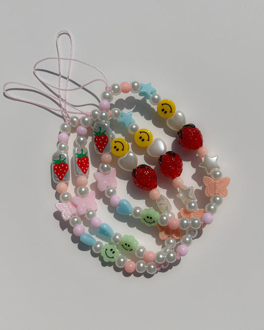 A studio shot of multiple Buttercup Studio Strawberry Jam Phone Straps. Made with pearls, colourful beads, white heart and star beads, a pink butterfly bead, two smiley face beads, a glass hand painted rectangle bead with a strawberry on it and a lampwork glass strawberry bead.