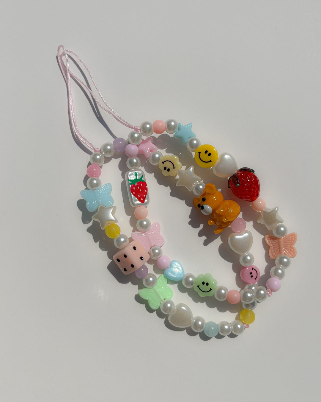 A studio shot of two Buttercup Studio Strawberry Jam Phone Straps. Made with pearls, colourful beads, white heart and star beads, a pink butterfly bead, two smiley face beads, a glass hand painted rectangle bead with a strawberry on it and a lampwork glass strawberry bead.