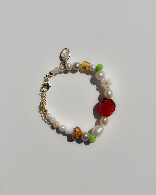 A studio shot of Buttercup Studio's Summertime Strawberry Freshwater Pearls Bracelet. Features assorted freshwater pearls, green and amber beads, a white daisy bead, a brown lampwork mushroom glass bead and a lampwork glass strawberry bead. 