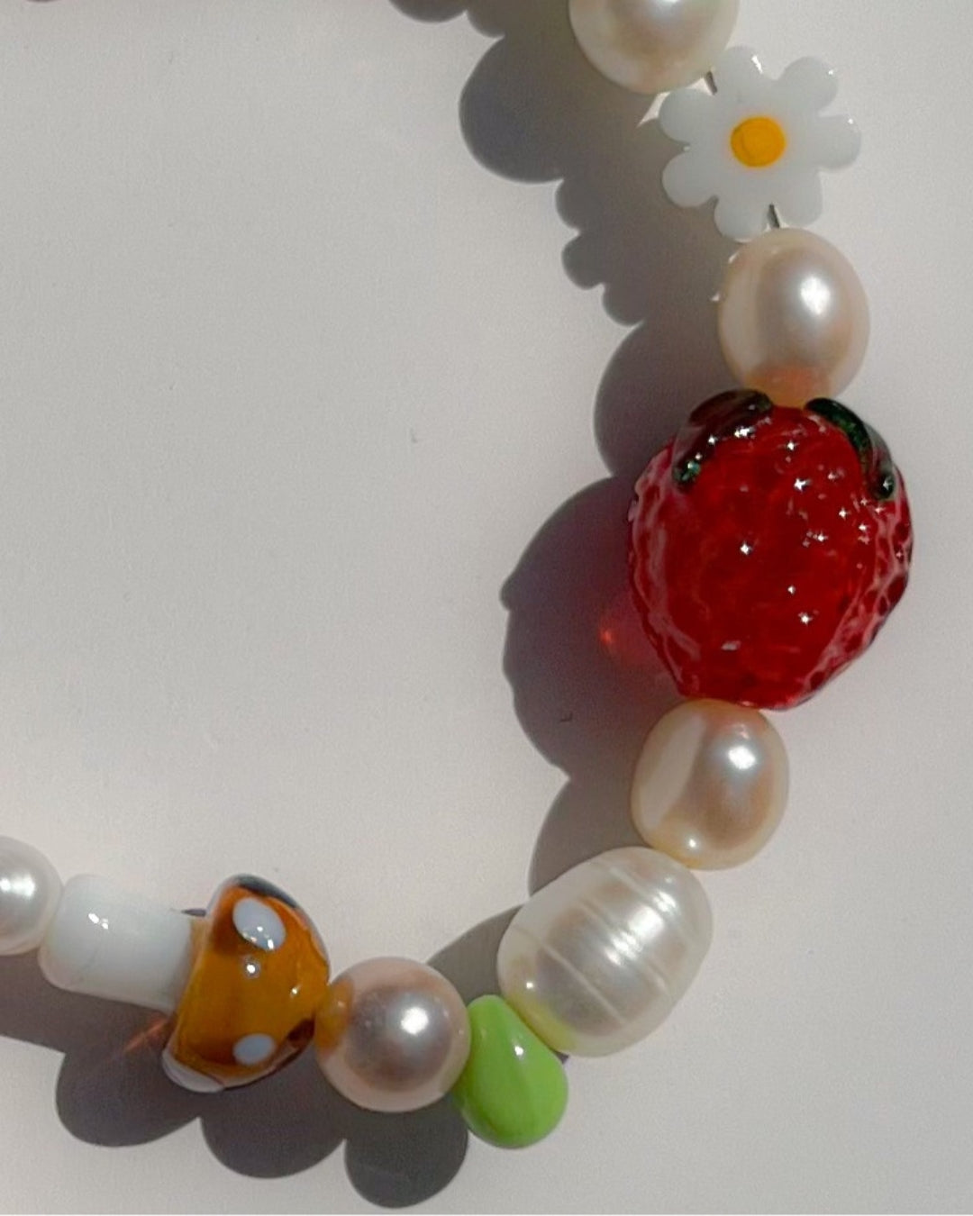 A close up studio shot of Buttercup Studio's Summertime Strawberry Freshwater Pearls Bracelet. Features assorted freshwater pearls, green and amber beads, a white daisy bead, a brown lampwork mushroom glass bead and a lampwork glass strawberry bead.