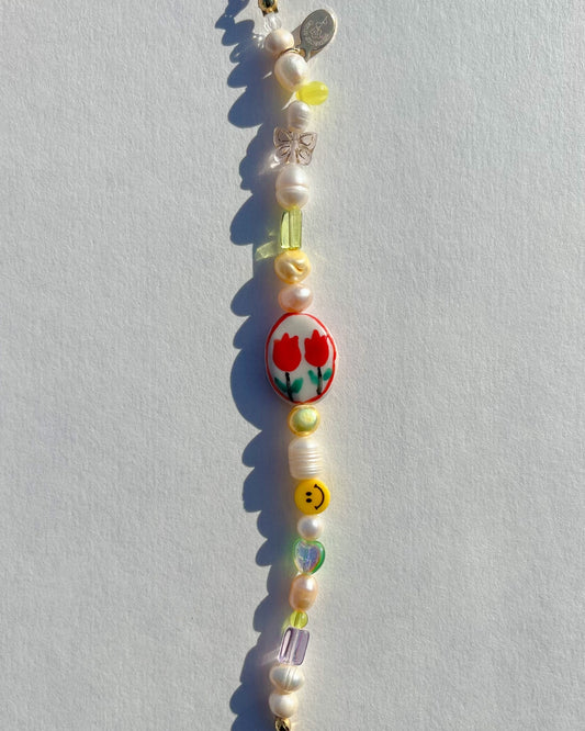 A studio shot of Buttercup Studio's Summertime Freshwater Pearls Bracelet. Made with assorted freshwater pearls, yellow, purple and green beads, a smiley face bead and a special lampwork hand painted glass rectangle bead with two red tulips on it. 