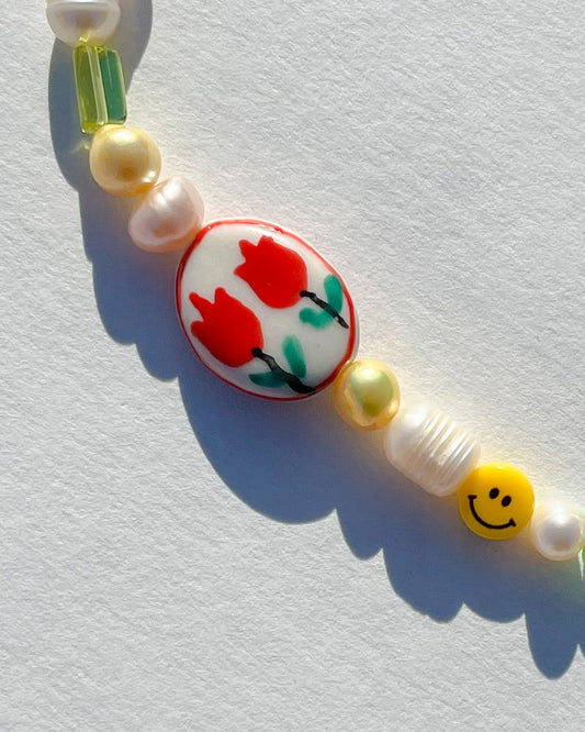 A close up shot of Buttercup Studio's Summertime Freshwater Pearls Bracelet. Made with assorted freshwater pearls, yellow, purple and green beads, a smiley face bead and a special lampwork hand painted glass rectangle bead with two red tulips on it. 