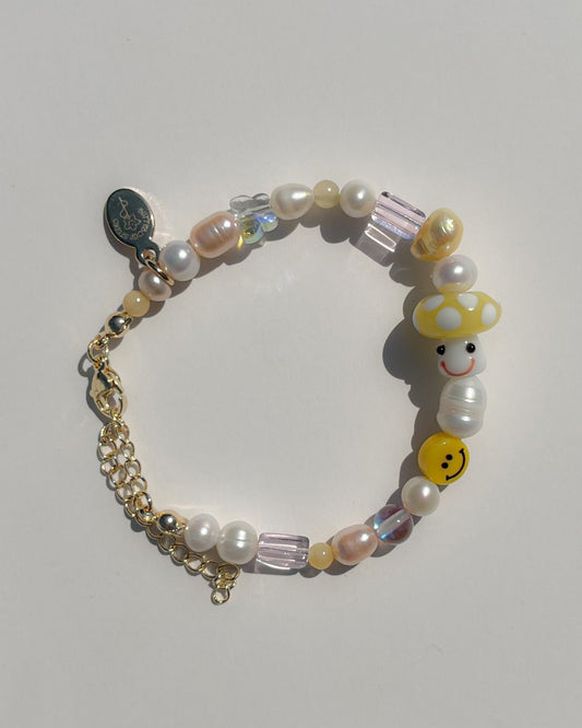 A studio shot of the Buttercup Studio Yellow Shroomie Freshwater Pearls Bracelet. Made with assorted freshwater pearls, clear and pink beads, a yellow smiley face bead and a special lampwork glass yellow mushroom bead. 