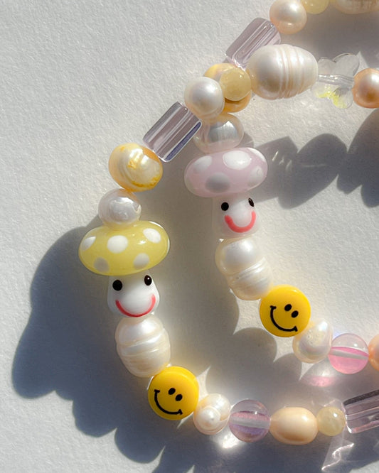 A studio shot of two Buttercup Studio Yellow Shroomie Freshwater Pearls Bracelets. Made with assorted freshwater pearls, clear and pink beads, a yellow smiley face bead and a special lampwork glass yellow mushroom bead.
