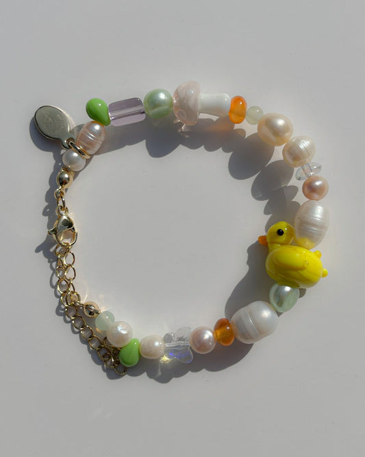 A studio shot of Buttercup Studio's Wonderland Duckie Freshwater Pearls Bracelet. Includes assorted freshwater pearls, orange and green beads, a lampwork glass pink mushroom bead and a yellow duck lampwork glass bead. 