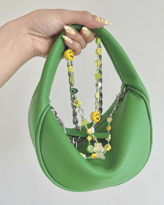 A studio shot of a hand holding up a mini green shoulder bag with a Buttercup Studio Custom Multipurpose Strap made with green and yellow beads and charms, attached to it. 