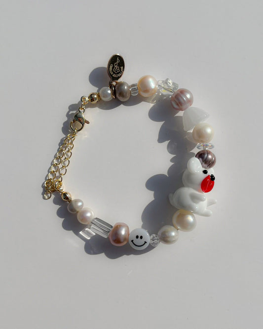 A studio shot of the one of one Angel Teddy Freshwater Pearls Bracelet by Buttercup Studio. Handmade using high-quality clear beads, gemstones, a white smiley face charm, locally sourced freshwater pearls in white, pink, cream, and grey, and a lampwork glass bead featuring a white teddy with a red nose. Made with an adjustable chain, 14k gold clasps and a Buttercup Studio emblem.