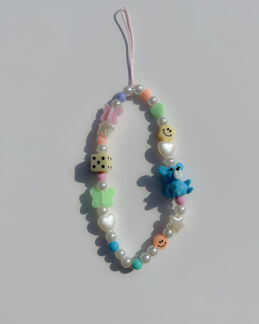A studio shot of the Aqua Confetti Blue Teddy personalized phone charm by Buttercup Studio. Hand-threaded using pink nylon with colourful acrylic beads, a pink butterfly and green butterfly bead, white stars and hearts beads, a coral smiley face and pastel yellow smiley face bead, and pearls. Features a glass blue teddy lampwork bead.