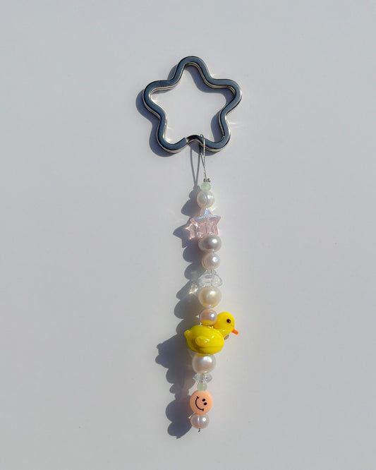 A studio shot of the one of one Baby Duckling Freshwater Pearls Lucky Charm keychain. Handmade using stainless steel wire looped on a star shaped keyring, this freshwater pearl keychain features white and pink pearls, clear beads, a sheer pink star bead, a coral smiley face bead and a yellow baby duck lampwork glass bead. 