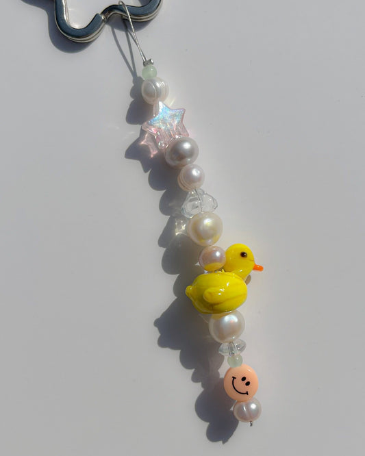 A close up studio shot of the one of one Baby Duckling Freshwater Pearls Lucky Charm keychain. Handmade using stainless steel wire looped on a star shaped keyring, this freshwater pearl keychain features white and pink pearls, clear beads, a sheer pink star bead, a coral smiley face bead and a yellow baby duck lampwork glass bead. 