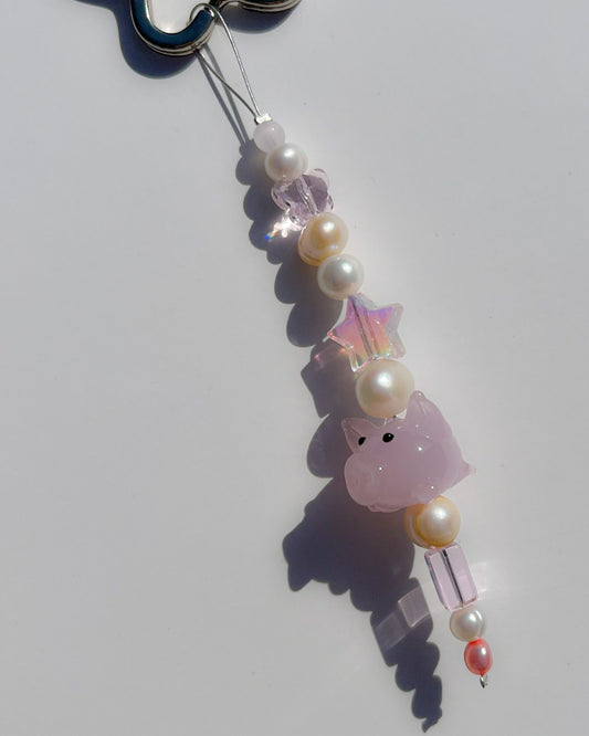 A close up studio shot of the one of one Baby Piglet Freshwater Pearls Lucky Charm keychain. Handmade using stainless steel wire looped on a star shaped keyring, this freshwater pearl keychain features white and yellow pearls, clear beads, sheer pink beads and a pink baby pig lampwork glass bead. 