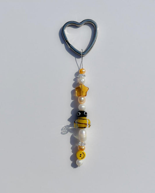 A studio shot of the one of one Busy Bee Freshwater Pearls Lucky Charm keychain. Handmade using stainless steel wire looped on a heart shaped keyring, this freshwater pearl keychain features white and yellow pearls, clear beads, a sheer orange star bead, a yellow smiley face bead and a yellow bee lampwork glass bead. 