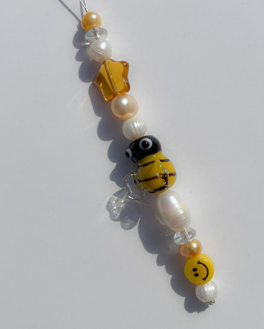 A close up studio shot of the one of one Busy Bee Freshwater Pearls Lucky Charm keychain. Handmade using stainless steel wire looped on a heart shaped keyring, this freshwater pearl keychain features white and yellow pearls, clear beads, a sheer orange star bead, a yellow smiley face bead and a yellow bee lampwork glass bead. 