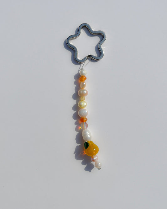A studio shot of the one of one Citrine Amber Pear Freshwater Pearls Lucky Charm keychain. Handmade using stainless steel wire looped on a star shaped keyring, this freshwater pearl keychain features white, pink and yellow pearls, sheer orange beads, and a yellow pear lampwork glass bead. 
