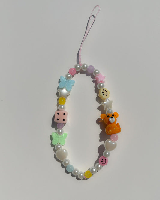 A studio shot of the Clementine Teddy Confetti personalized phone charm by Buttercup Studio. Hand-threaded using pink nylon with colourful acrylic beads, a blue butterfly and green butterfly bead, white stars and hearts beads, a pink smiley face and pastel yellow smiley face bead, and pearls. Features an orange teddy lampwork glass bead.