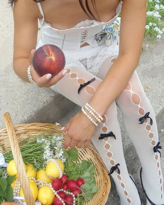A close up shot of a model sitting on concrete steps, wearing a white spaghetti strap corset and white netted stockings with black bows throughout. They are holding a peach in one hand while the other hand is reaching for a woven picnic basket filled with flowers, lemons, radishes, herbs and strong pearls. The model wears Buttercup Studio's Timeless Pastel Freshwater Pearls Bracelet on their wrists.