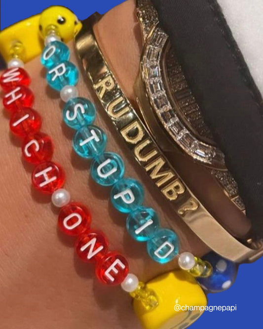 A close up of Buttercup Studio's Aubrey Set on Drake's wrist. A gold curved bracelet band carved with the phrase R U DUMB? with the Drake Custom Bracelet set in teal beads spelling out OR STUPID below it, and a Drake Custom Bracelet in red beads spelling out WHICH ONE at the bottom of the set.   Image credits: @champagnepapi on Instagram.