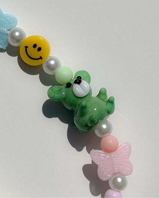 Close up studio shot of Buttercup Studio's Emerald Teddy Confetti Phone Strap. Featuring a special teddy handmade lampwork glass bead, smiley face bead, pink butterfly bead, acrylic pink beads and pearls.