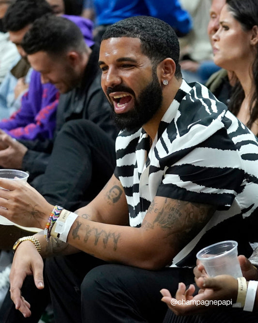 Drake seated at a sports event wearing a custom Drake bracelet from the Aubrey set by Buttercup Studio. Image credits @champagnepapi on Instagram.