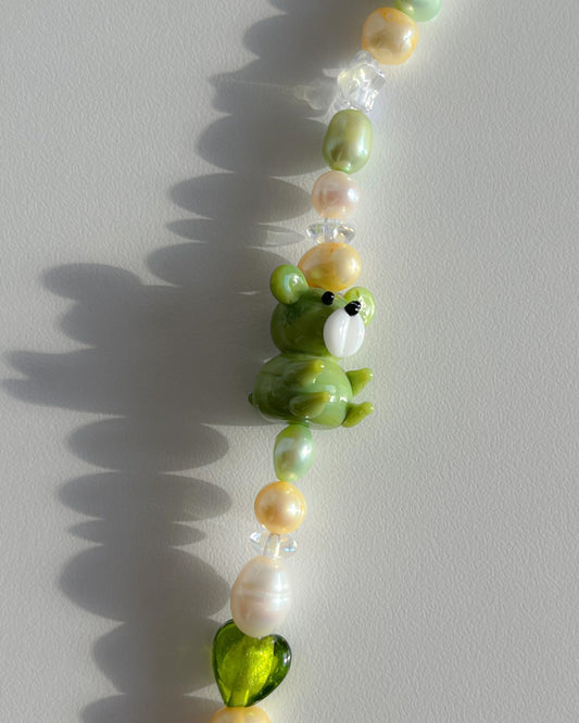 A close up studio shot of Buttercup Studio's Forest Teddy Freshwater Pearls Bracelet. Features clear beads, a sheer green heart bead, assorted yellow, green and white freshwater pearls, and a special green teddy lampwork glass bead.