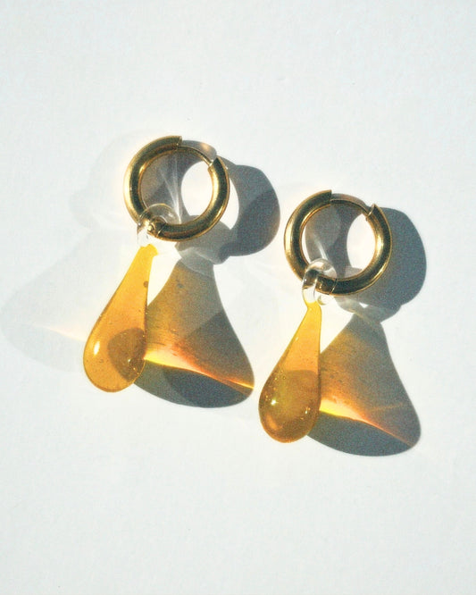 A studio shot of the Celeste earrings in honey from Buttercup Studio. The Celeste Earrings are made with thick 18k gold plated tarnish-resistant, hypoallergenic 12 mm thick hoops and high-quality 22mm lampwork glass water drop pendants.