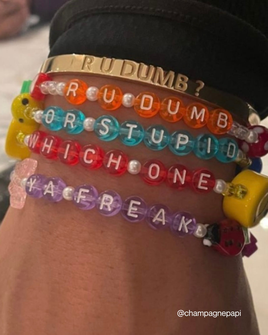 A close up of Buttercup Studio's Aubrey Set on Drake's wrist. A gold curved bracelet band carved with the phrase R U DUMB? with the Drake Custom Bracelet set in orange beads spelling out R U DUMB? below it, and a Drake custom bracelet set in teal beads spelling out OR STUPID below that, and a Drake Custom Bracelet in red beads spelling out WHICH ONE under it, and a Drake Custom Bracelet set in purple beads spelling out YA FREAK at the bottom. Image credits: @champagnepapi on Instagram.