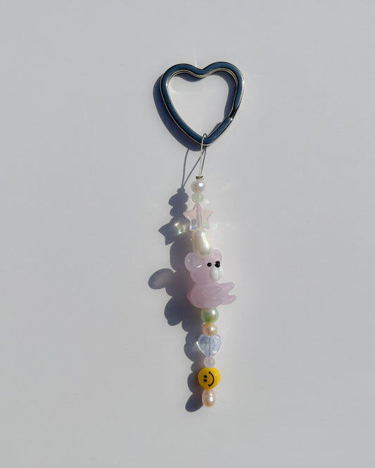 A studio shot of the one of one Garden Teddy Freshwater Pearl Lucky Charm keychain. Handmade using stainless steel wire looped on a heart shaped keyring, this freshwater pearl keychain features a yellow smiley charm, clear star and heart beads, assorted white, pink and green freshwater pearls and a special pink teddy lampwork glass bead.