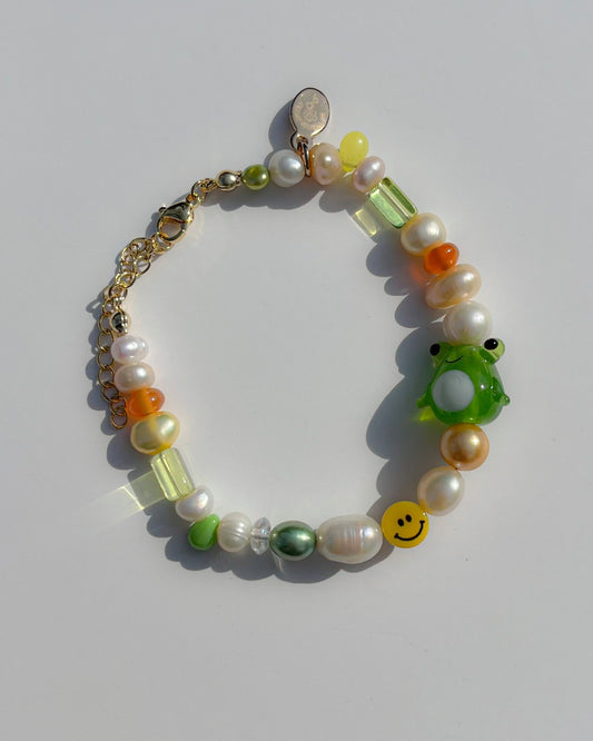 A studio shot of Buttercup Studio's Happy Froggie Freshwater Pearls Bracelet. Made with assorted freshwater pearls, sheer amber and green beads, a yellow smiley face bead and a special green frog lampwork glass bead.