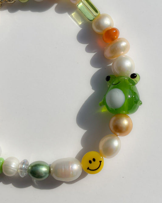 A close up studio shot of Buttercup Studio's Happy Froggie Freshwater Pearls Bracelet. Made with assorted freshwater pearls, sheer amber and green beads, a yellow smiley face bead and a special green frog lampwork glass bead.