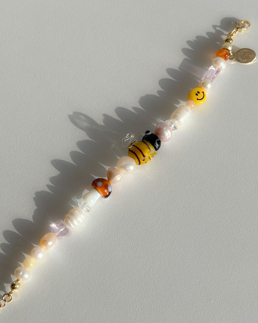 A studio shot of Buttercup Studio's Honeycomb Bee Freshwater Pearls Bracelet. Made with assorted freshwater pearls, clear pink and amber beads, a yellow smiley face bead, a special brown mushroom lampwork glass bead and a special yellow bee lampwork glass bead. 