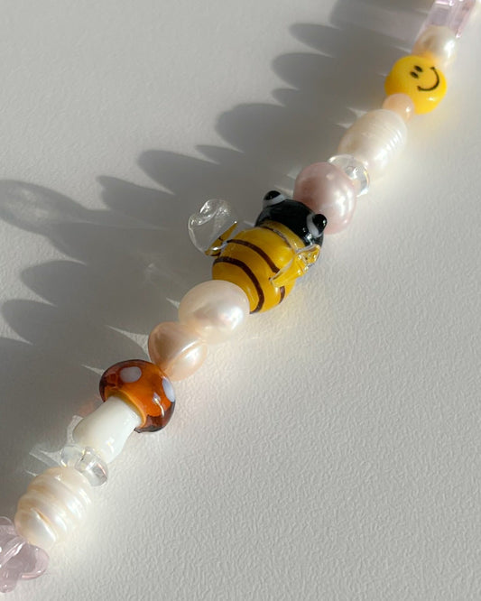 A close up studio shot of Buttercup Studio's Honeycomb Bee Freshwater Pearls Bracelet. Made with assorted freshwater pearls, clear pink and amber beads, a yellow smiley face bead, a special brown mushroom lampwork glass bead and a special yellow bee lampwork glass bead.