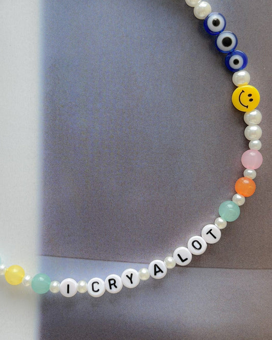 A close up studio shot of the Buttercup Studio Custom Regular Pearls Necklace, spelling out "I CRY A LOT", made exclusively for happytears.ca. Includes regular pearls, colourful teal, pink and orange beads, a yellow smiley face bead and three evil eye beads.