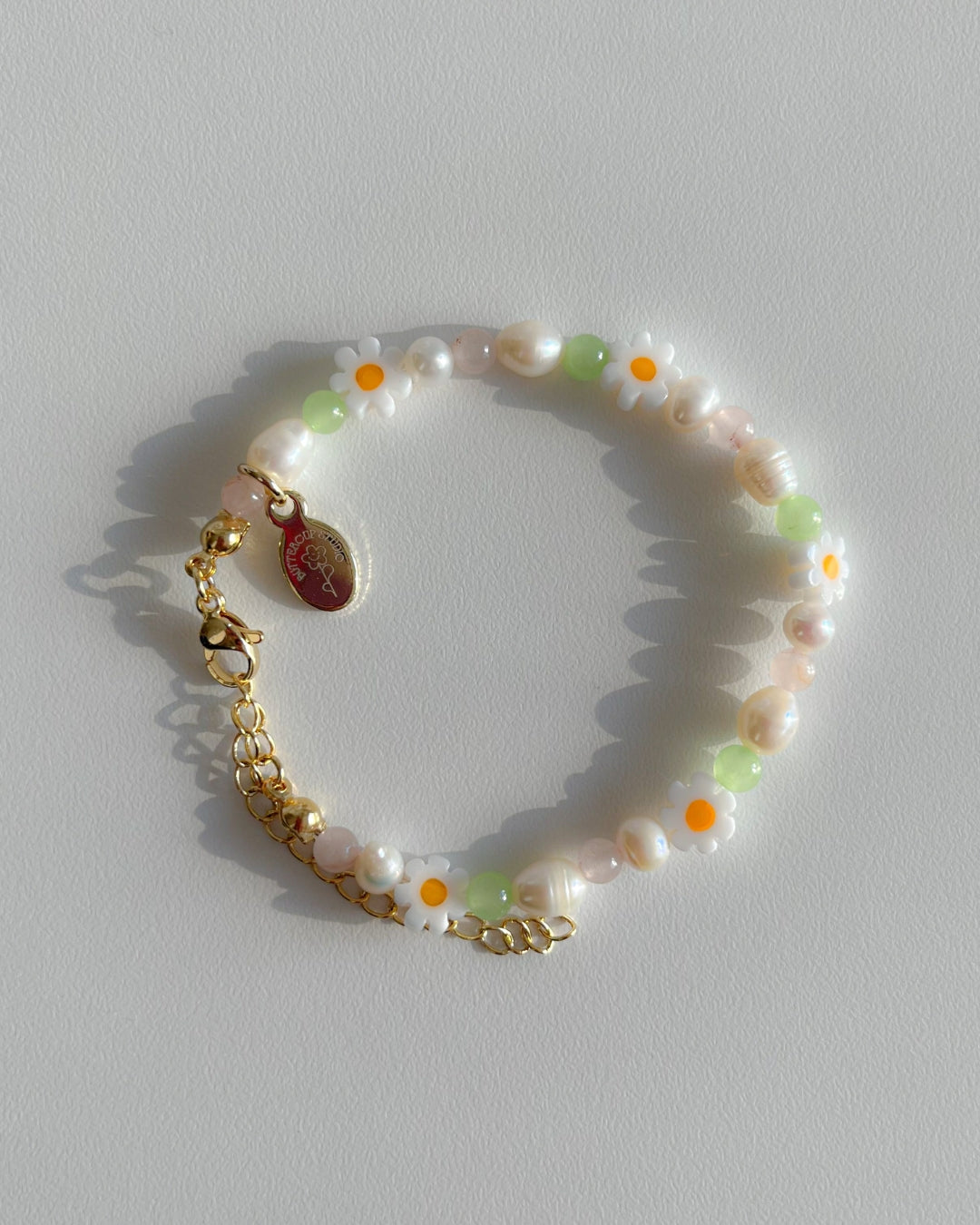 Close up studio shot of multiple Daisy For You Freshwater Pearls Bracelets by Buttercup Studio. Features freshwater pearls in assorted colours and sizes, green beads and daisy charms.